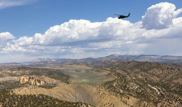 A helicopter with a long cable load flying over scenic desert landscape