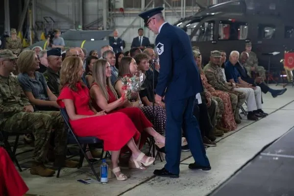 A uniformed servicemember hands a bouquet of red roses to a woman in a red gown