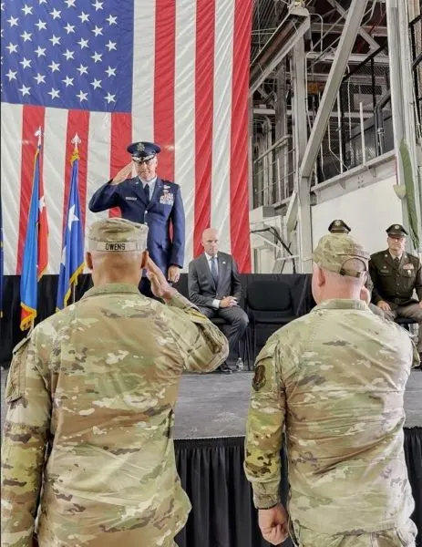 Two servicemembers in camouflage salute a superior officer in a dress uniform