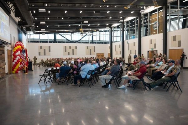 Audience of civilians and military members seated in rows of chairs listening to an indoor presentation.