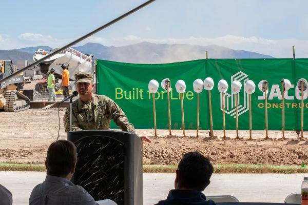 Military officer speaking at a groundbreaking event with white hardhats on shovels in front of a green construction banner.