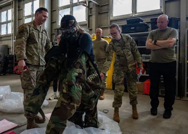 An airman puts on a camouflage MOPP suit inside a hangar while military instructors look on during training