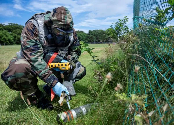 An airman in full MOPP gear crouches in the grass to inspect an unexploded ordnance casing with detection equipment