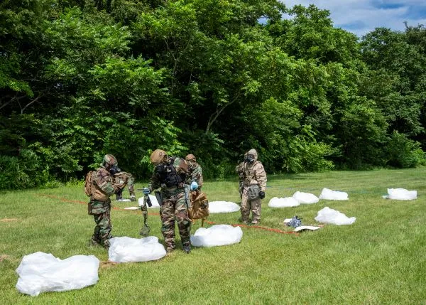 Military personnel in full CBRN protective suits line up near large white plastic bags during an outdoor training exercise on a grassy field