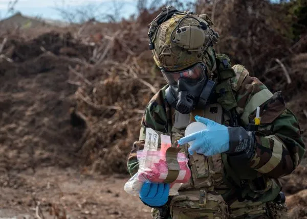 An airman in full MOPP gear and gas mask holds open a clear plastic emergency response kit while wearing blue protective gloves
