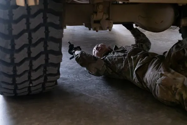 A soldier lying on their back underneath a military truck to inspect and repair the wheel axle assembly.