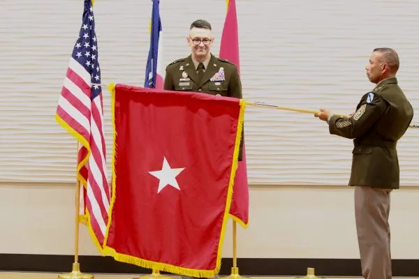 A horizontal view of an NCO presenting a red Brigadier General flag with a single white star alongside a newly promoted Army officer.