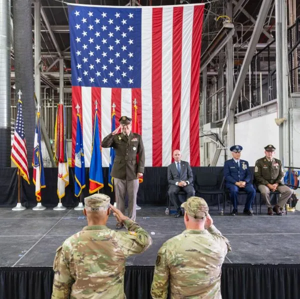 Two service members in camouflage salute a military officer standing on stage.
