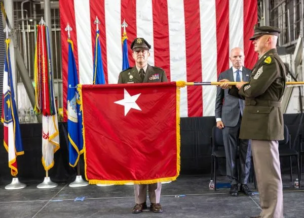 An Army officer stands on stage beside a newly unfurled red one-star general officer flag.