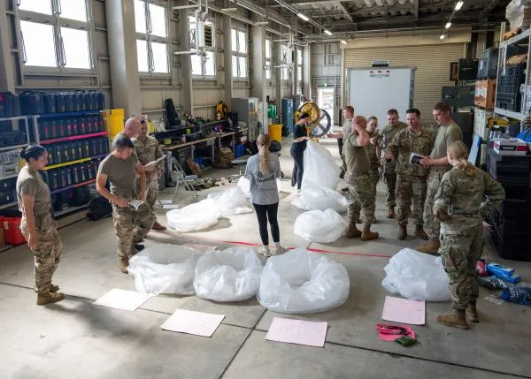 Military personnel gather inside an equipment facility around large white plastic bags arranged on the floor during training