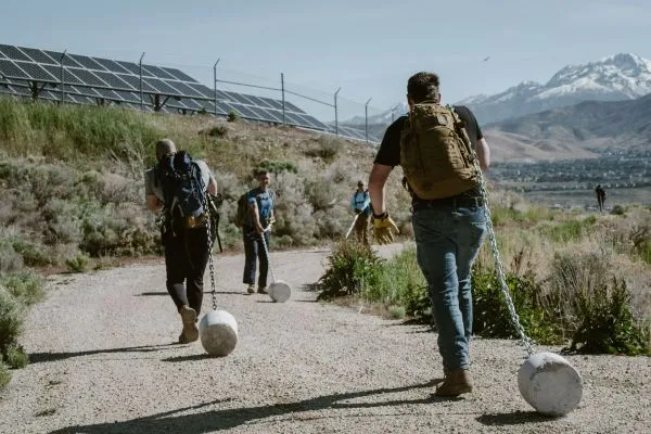 Three individuals carry cement blocks with heavy-duty silver chains along a path and dirt trail