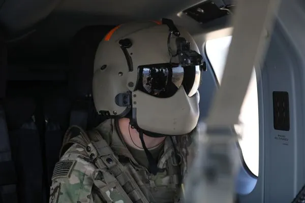 A U.S. military helicopter pilot wearing a full flight helmet with a dark visor seated inside an aircraft cabin.