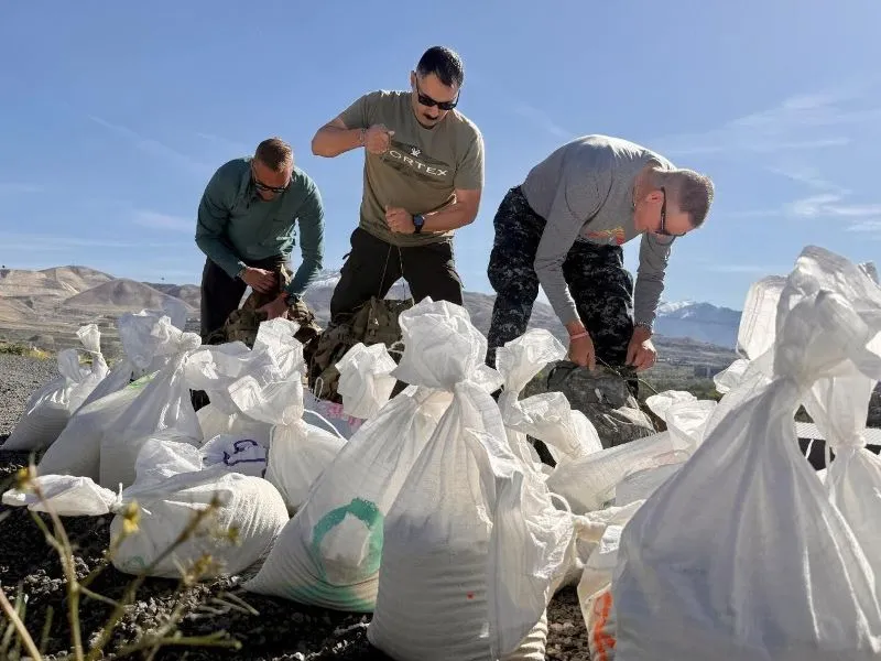 Three individuals tie ruck sacks behind a pile of white sandbags