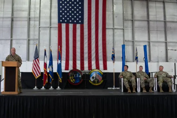 An Air Force speaker at a podium addressing seated military leadership on stage under a large American flag inside a hangar