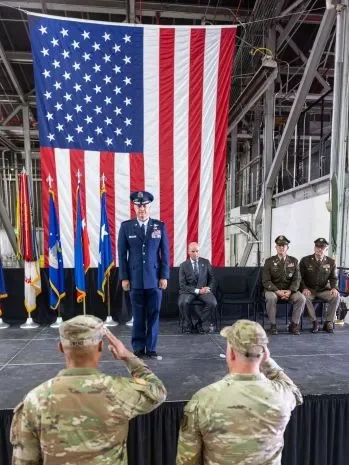 Two service members in camouflage salute a military officer standing on stage.