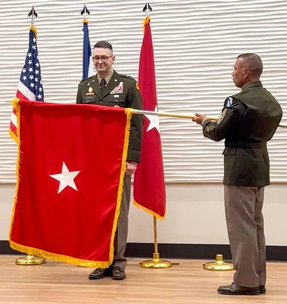 A U.S. Army NCO unfurls a red single-star Brigadier General flag in front of a newly promoted general officer during a ceremony.