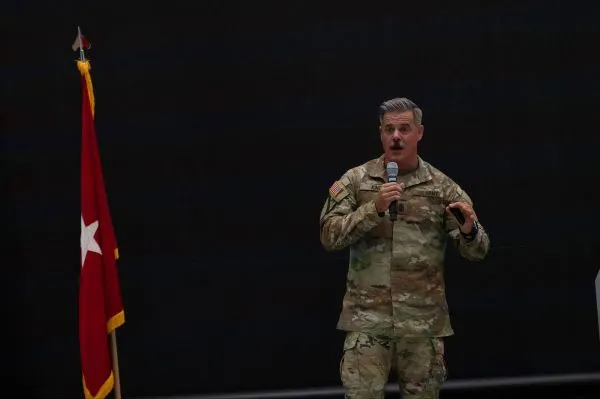 A U.S. Army officer in camouflage uniform named King holding a microphone and gesturing while speaking on stage next to a red flag with a white star.