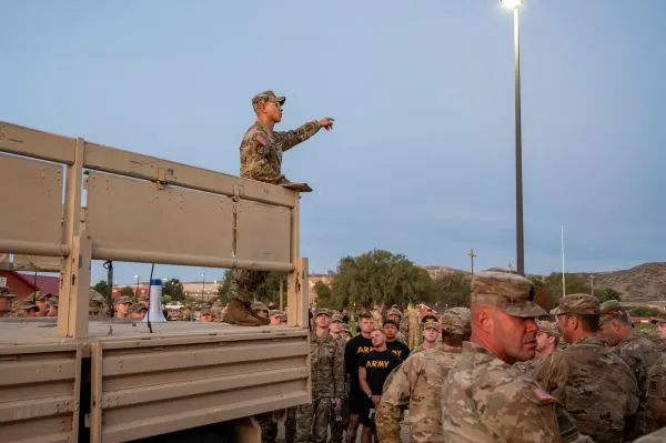 Military instructor gesturing from a elevated vehicle bed to a group of assembled Soldiers